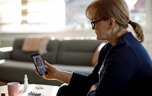 A woman with glasses sitting on a couch while holding a smartphone and video chatting with her concierge medicine doctor